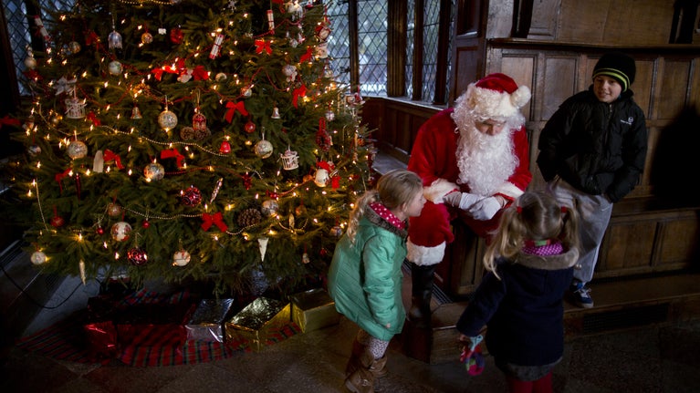 Santa Claus seated beside a Christmas tree, interacts with two young girls in coats, while a young boy looks on. The tree is decorated with lights, bows, and ornaments, with wrapped gifts at its base. The scene takes place in a warmly lit, paneled room.